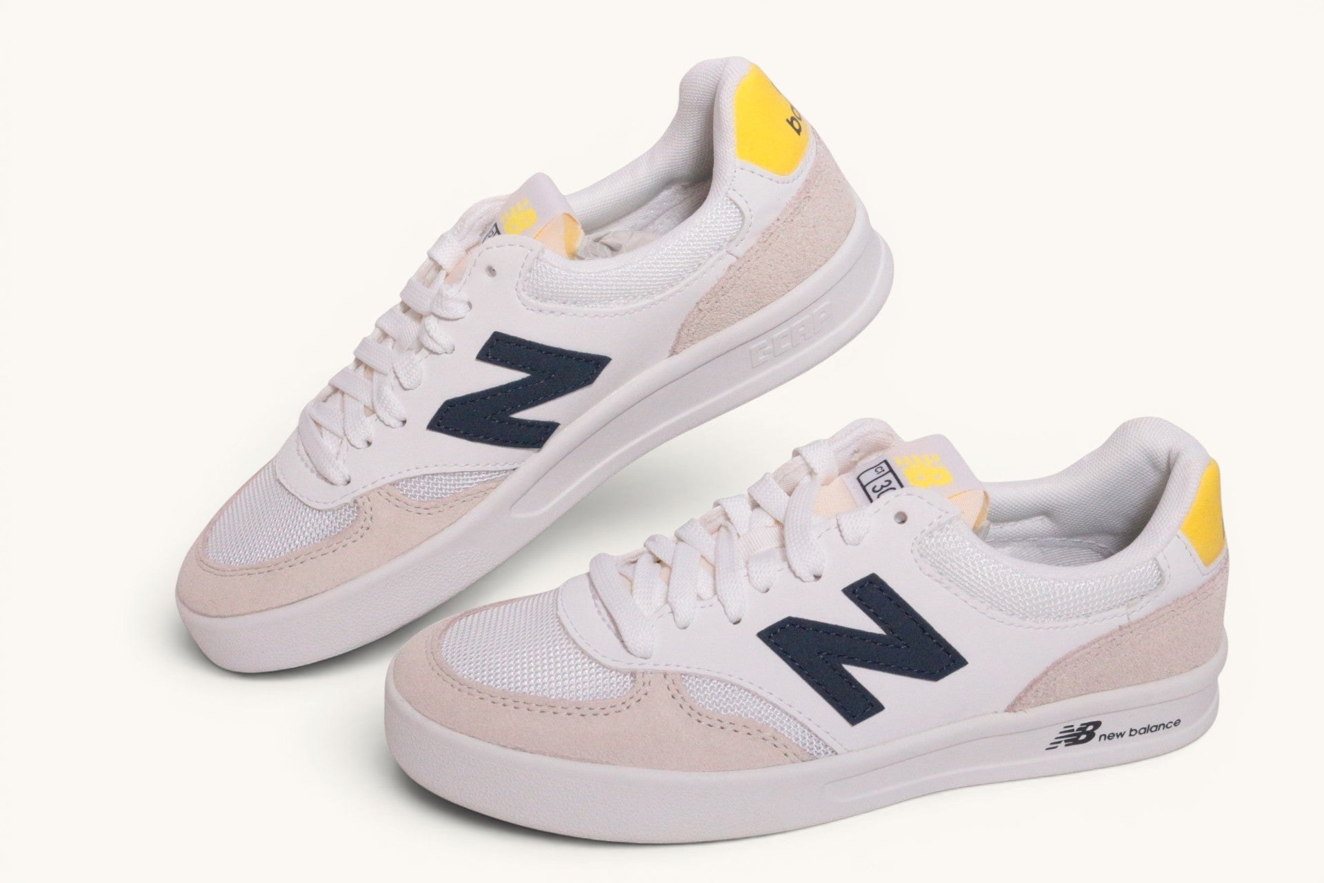 Baskets New Balance Frippindealz blanches en cuir et mesh avec logo bleu marine et détails jaunes, style unisexe.