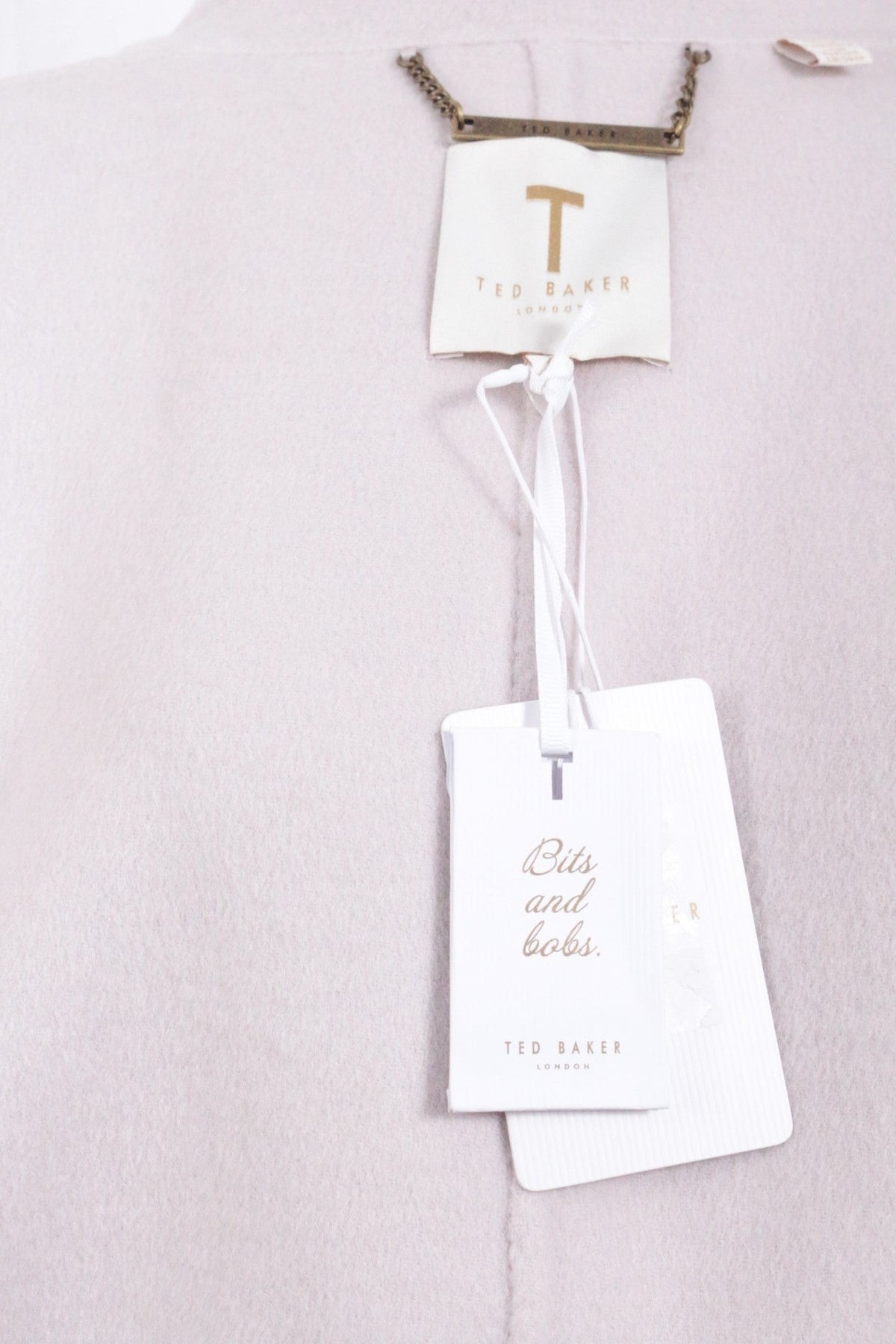 Gros plan sur l'étiquette et la chaîne dorée d'un manteau en laine beige Ted Baker neuf chez Frippindealz